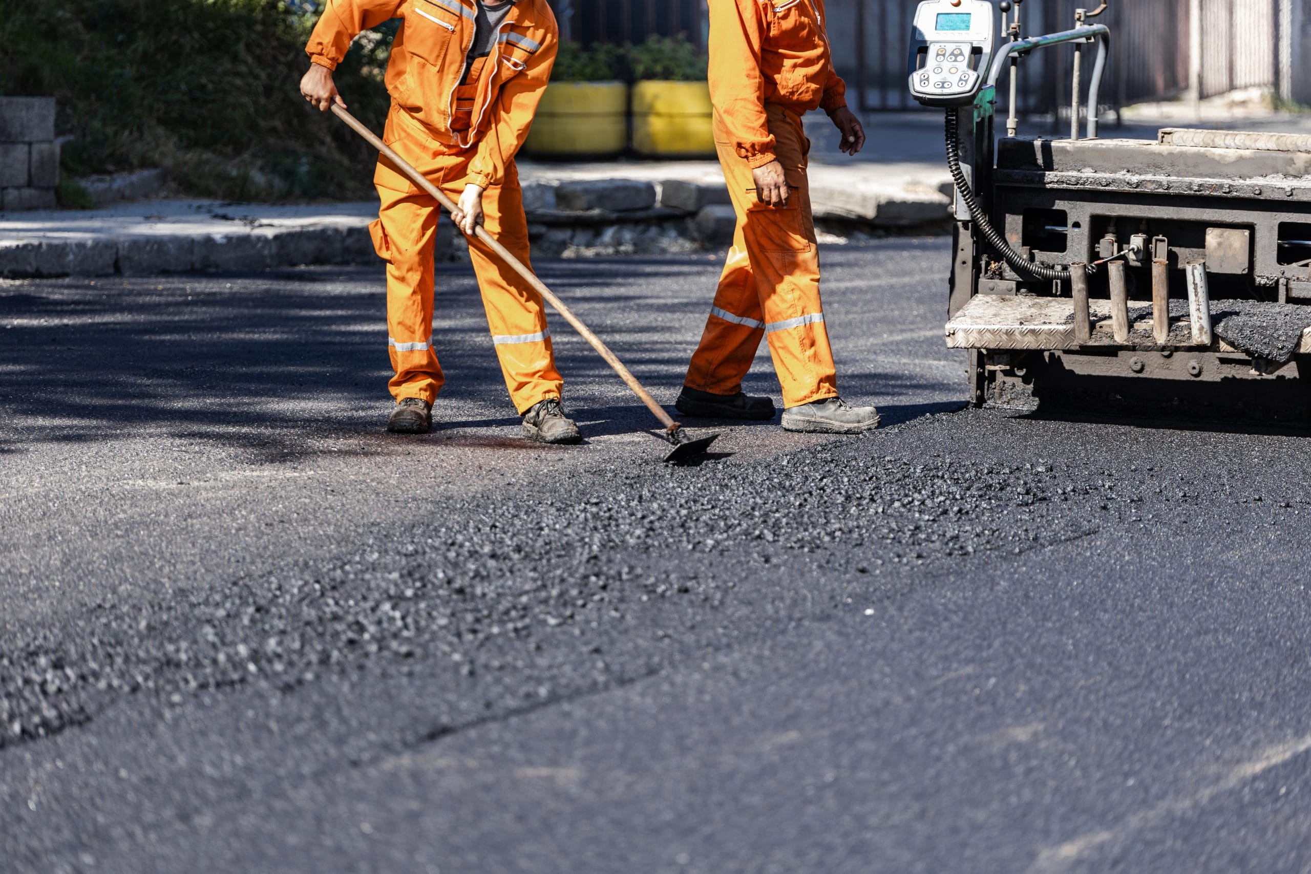 Asphalt Contractors Two workers in orange uniforms spreading asphalt on a road beside a paving machine.