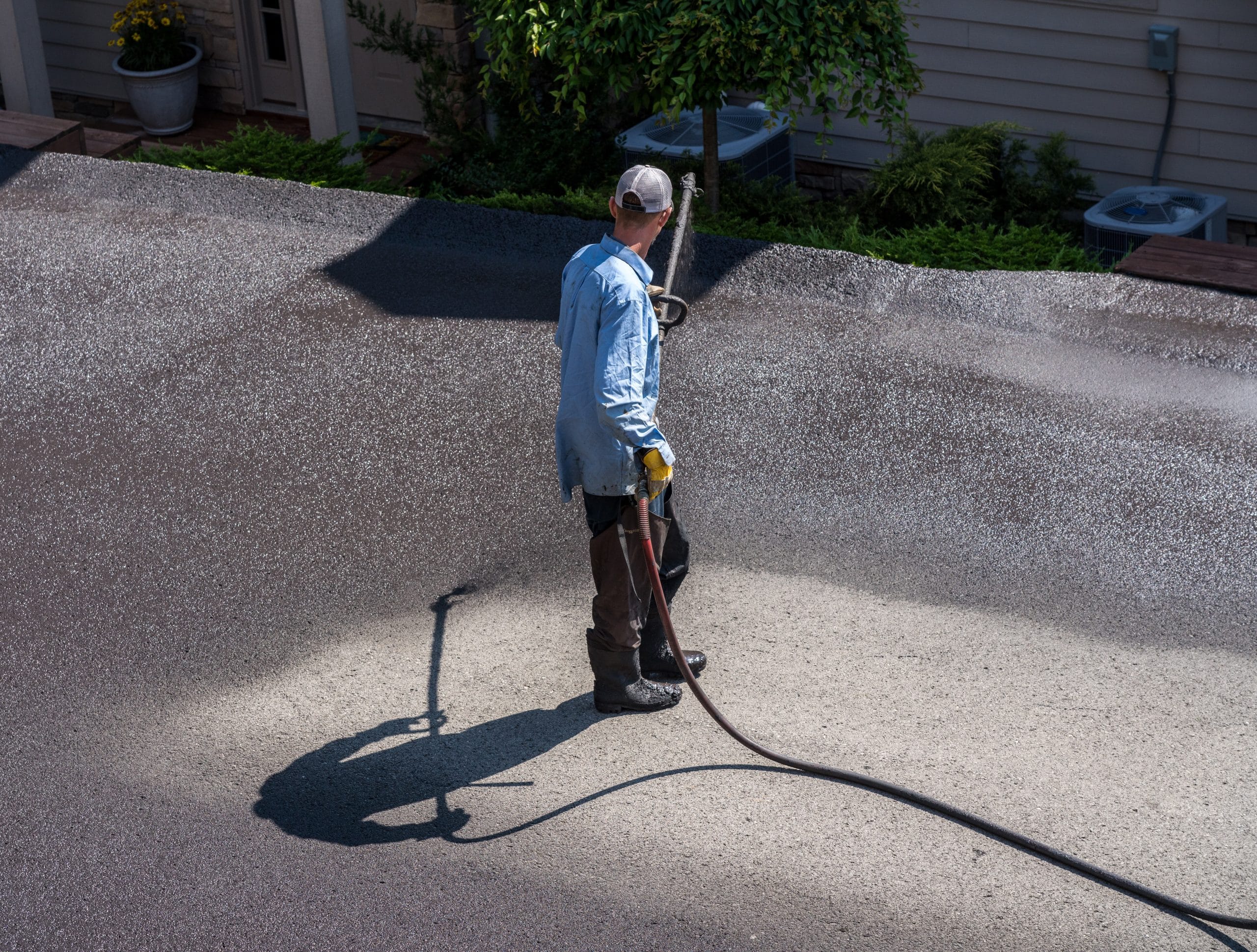 Person spraying sealant on flat roof with hose, wearing blue shirt, cap, and boots.