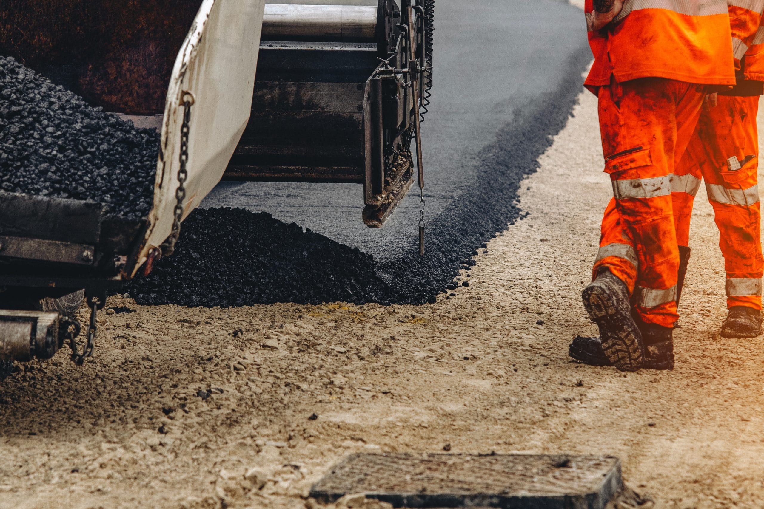 Road construction: Worker in orange gear standing by machine laying asphalt on a road.