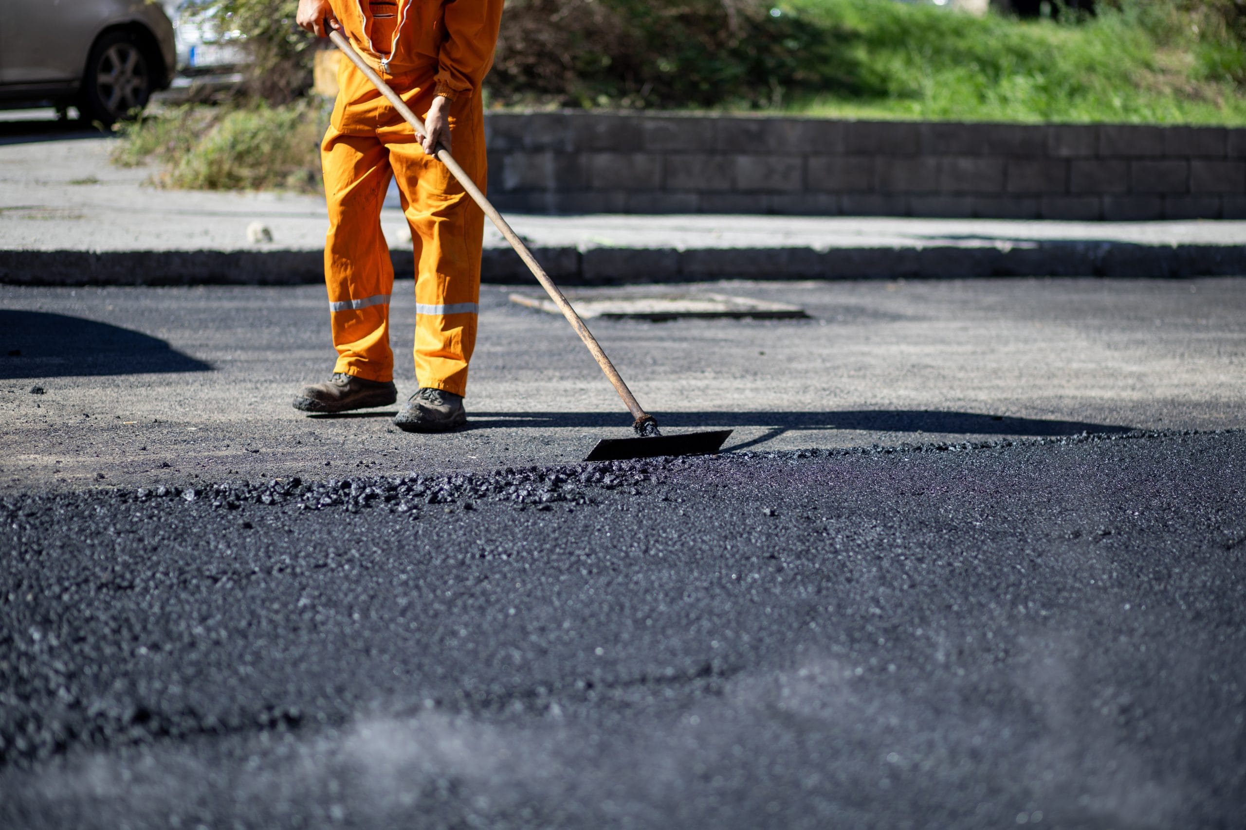 Worker in orange uniform spreads fresh asphalt on a road with a rake under daylight.